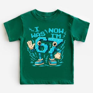 A green t-shirt with a graphic design featuring a robot character and text that says "I WAS NOW I'M 67".