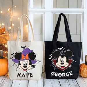 Peut inclure: Deux sacs fourre-tout sur le thème d'Halloween. L'un est de couleur crème avec un motif de Minnie Mouse sorcière et le nom "KATE". L'autre est noir avec un motif de Mickey Mouse vampire et le nom "GEORGE". Les deux sacs ont des anses noires.