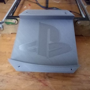 May include: A gray, 3D-printed PlayStation logo. The logo is embossed on a rectangular base with curved edges and mounting holes. The item appears to be a component or accessory, possibly for a gaming setup.