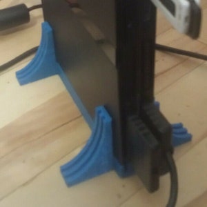 May include: Black PlayStation 2 console with a green power light, supported by a blue stand. A USB drive is plugged in, also with a green light. Cables are connected to the console.