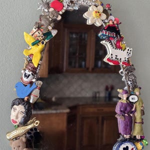 K&ouml;nnte beinhalten: Ovaler Spiegel mit einem dekorativen Rand aus verschiedenen Charms, darunter eine Freiheitsstatue, ein Teddyb&auml;r, eine Katze und eine Bowlingkegel. Der Rand ist mit Strasssteinen und anderen kleinen Schmuckst&uuml;cken verziert. Der Spiegel reflektiert einen Raum.