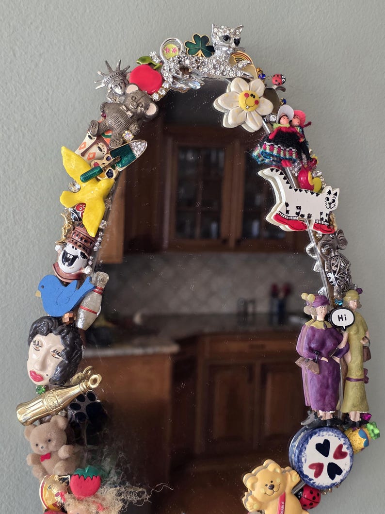 K&ouml;nnte beinhalten: Ovaler Spiegel mit einem dekorativen Rand aus verschiedenen Charms, darunter eine Freiheitsstatue, ein Teddyb&auml;r, eine Katze und eine Bowlingkegel. Der Rand ist mit Strasssteinen und anderen kleinen Schmuckst&uuml;cken verziert. Der Spiegel reflektiert einen Raum.