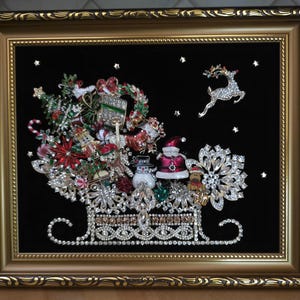 Könnte beinhalten: Ein gerahmtes Wandbild mit einem dekorativen Schlitten aus klaren Strasssteinen und Metall, gefüllt mit verschiedenen weihnachtlichen Broschen, darunter ein Schneemann, der Weihnachtsmann, ein Rentier und ein Baum, vor schwarzem Samthintergrund.