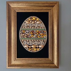 May include: A framed artwork featuring a jeweled egg design. The egg is adorned with various gold, amber, and green gemstones and rhinestones, set against a black background. The frame is a gold color.