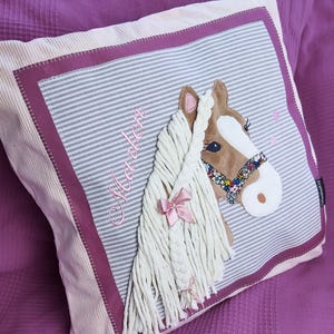 May include: A pink and white striped pillow with a brown and white horse head applique. The horse has a pink ribbon in its mane and a floral patterned bridle. The pillow is personalized with the name "Maddie" embroidered in pink.