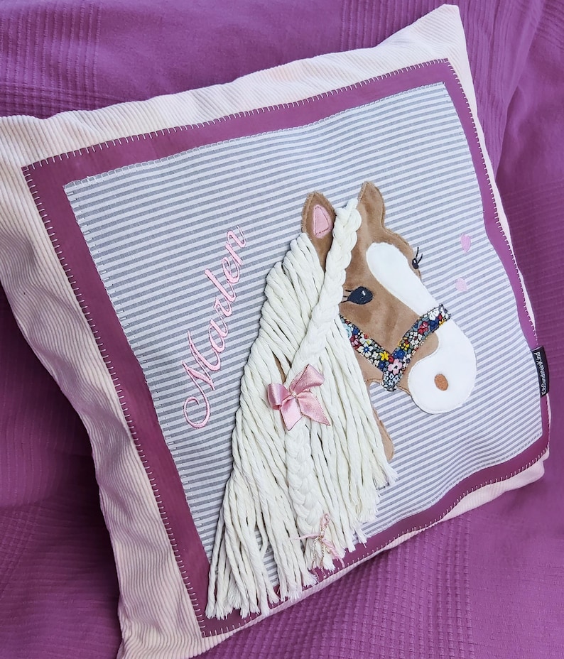 May include: A pink and white striped pillow with a brown and white horse head applique. The horse has a pink ribbon in its mane and a floral patterned bridle. The pillow is personalized with the name "Maddie" embroidered in pink.