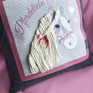 May include: A gray and white striped pillow with a pink border and a gray horse head applique. The horse has a white mane and tail, a pink ribbon, and a floral patterned bridle. The pillow has the word "Pferdeliebe" embroidered on it in pink.