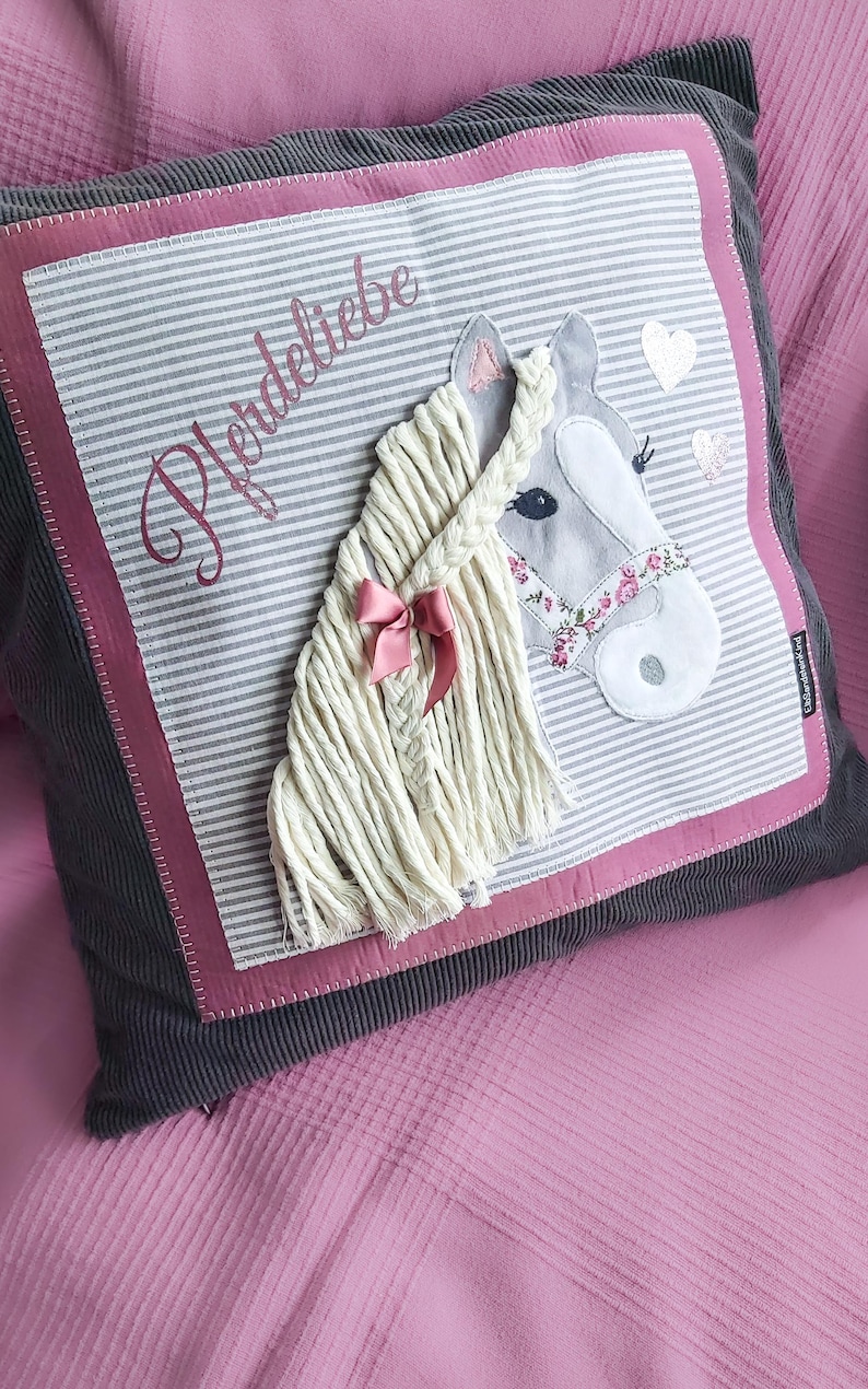 May include: A gray and white striped pillow with a pink border and a gray horse head applique. The horse has a white mane and tail, a pink ribbon, and a floral patterned bridle. The pillow has the word "Pferdeliebe" embroidered on it in pink.
