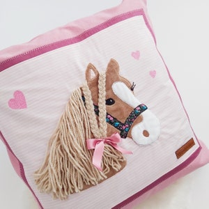 May include: Pink pillow with a brown horse applique with a pink ribbon and a braided mane. The pillow has a pink and white striped fabric with pink hearts.