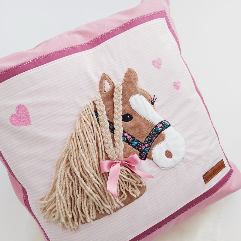 May include: Pink pillow with a brown horse applique with a pink ribbon and a braided mane. The pillow has a pink and white striped fabric with pink hearts.