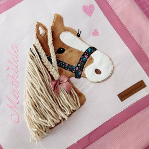 May include: A pink and white fabric tote bag with a brown horse applique. The horse has a braided mane and a pink bow. The bag has the name "Mathilda" embroidered on it and a brown leather tag that says "ElbSandsteinKind".