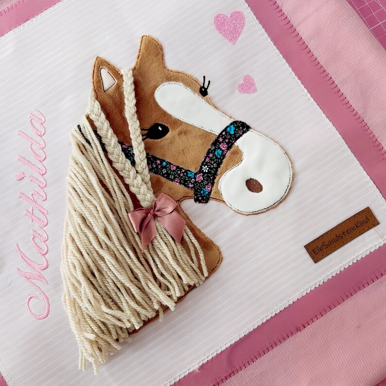 May include: A pink and white fabric tote bag with a brown horse applique. The horse has a braided mane and a pink bow. The bag has the name "Mathilda" embroidered on it and a brown leather tag that says "ElbSandsteinKind".