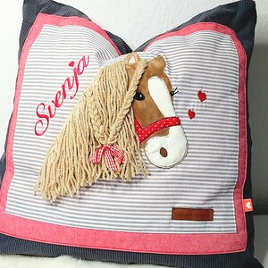 May include: A gray and pink pillow with a horse head applique. The horse has a brown face, white muzzle, and a long blonde mane. The pillow has the name "Svenja" embroidered on it in red thread. The pillow has a small red heart in the bottom right corner.