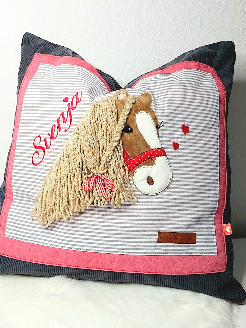 May include: A gray and pink pillow with a horse head applique. The horse has a brown face, white muzzle, and a long blonde mane. The pillow has the name "Svenja" embroidered on it in red thread. The pillow has a small red heart in the bottom right corner.