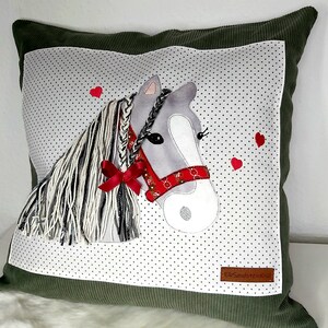 May include: A green pillow with a white background featuring a gray horse with a red ribbon and a red bandana. The horse has a braided mane and tail. The background is covered in small white dots. The pillow has a small brown label with the text "Elo Sandstein Kids".