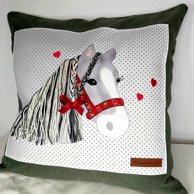May include: A green pillow with a white background featuring a gray horse with a red ribbon and a red bandana. The horse has a braided mane and tail. The background is covered in small white dots. The pillow has a small brown label with the text "Elo Sandstein Kids".