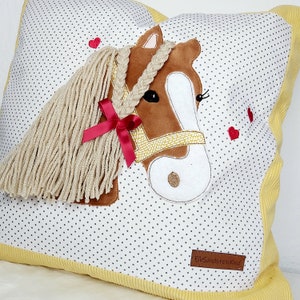 May include: A white pillow with a brown and white horse applique. The horse has a yellow braided mane and a red ribbon. The pillow has a yellow border and a small white label with the text "ElbSandsteinKind".