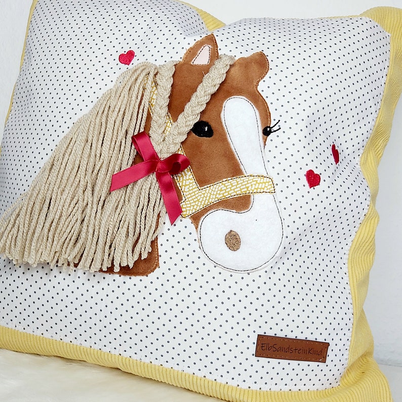 May include: A white pillow with a brown and white horse applique. The horse has a yellow braided mane and a red ribbon. The pillow has a yellow border and a small white label with the text "ElbSandsteinKind".