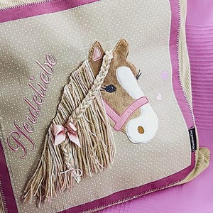 May include: A beige and pink pillow with a brown horse head applique. The horse has a pink bridle and a braided mane. The pillow has the word "Pferdeliebe" embroidered on it.
