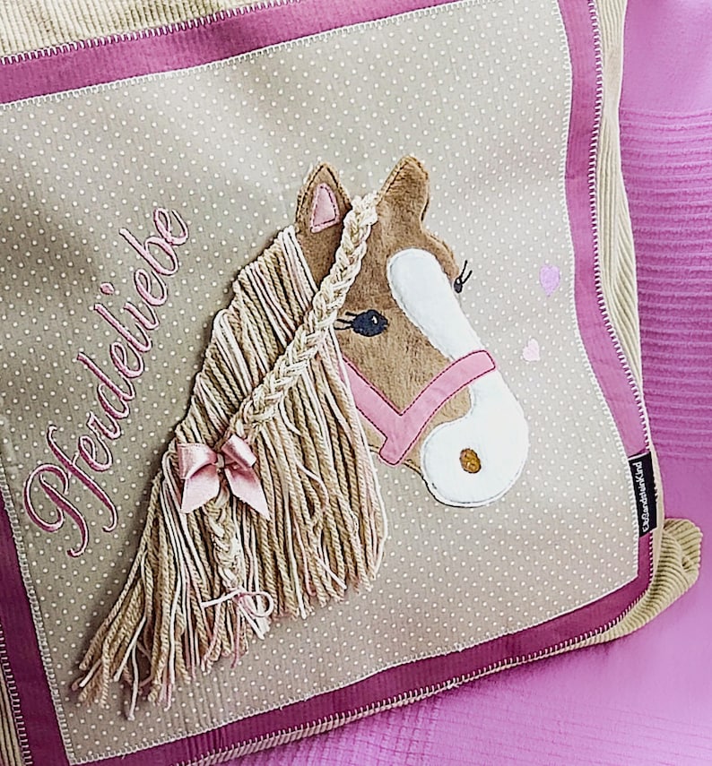 May include: A beige and pink pillow with a brown horse head applique. The horse has a pink bridle and a braided mane. The pillow has the word "Pferdeliebe" embroidered on it.