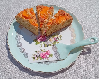 May include: A light blue and white floral patterned plate holds three slices of apricot cake. A matching cake server rests on the plate. The plate sits on a light gray tablecloth, with flowers in the background.