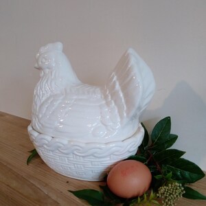 May include: A white ceramic hen-shaped container with a basket-weave base. The lid is a detailed hen. An egg and greenery are next to the container. The container is on a wooden surface.