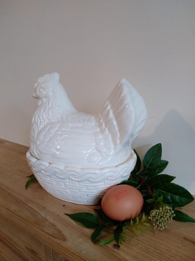 May include: A white ceramic hen-shaped container with a basket-weave base. The lid is a detailed hen. An egg and greenery are next to the container. The container is on a wooden surface.