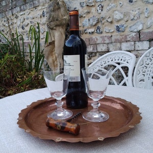 May include: A copper serving tray with a scalloped edge and decorative rivets. Two clear wine glasses and a bottle of red wine are on the tray. A wooden corkscrew is also on the tray.