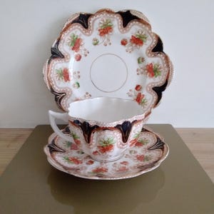 May include: A vintage teacup, saucer, and plate set. The white porcelain pieces feature a floral pattern with red, orange, and green accents. The edges are trimmed with a dark brown and black scalloped design. The set is displayed on a gold surface.