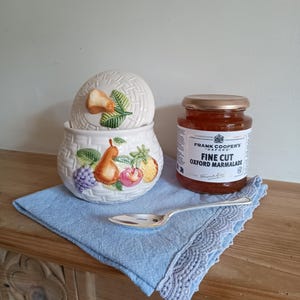 May include: A white ceramic jar with a lid decorated with fruit and a woven pattern. The jar is filled with marmalade. A jar of Frank Cooper's Fine Cut Oxford Marmalade is next to the jar. A silver spoon and a blue linen napkin are on a wooden surface.