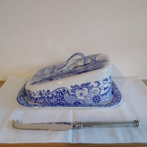 May include: A blue and white ceramic butter dish with a matching lid and a silver cheese knife. The dish features a floral and landscape design. The knife is approximately 20 cm long.