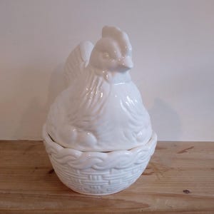 May include: A white ceramic hen-shaped container with a lid. The hen is perched atop a basket-weave base. The container is designed to hold eggs or other small items. The overall design is a classic, farmhouse style.