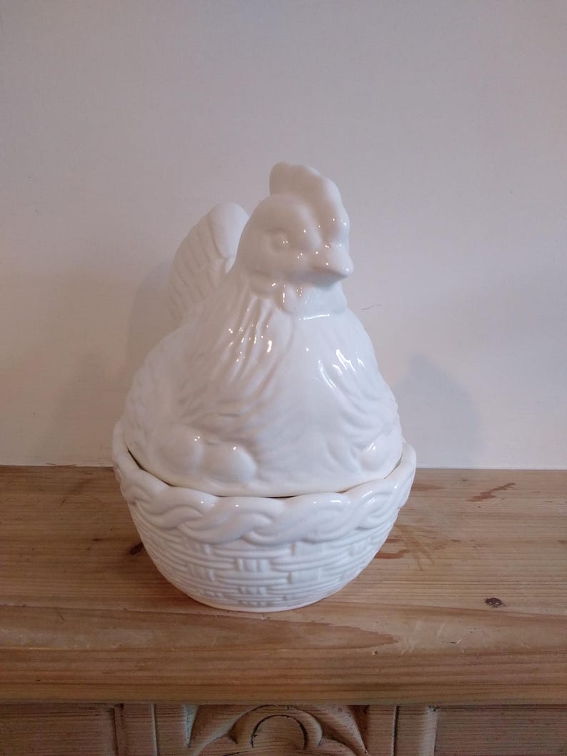 May include: A white ceramic hen-shaped container with a lid. The hen is perched atop a basket-weave base. The container is designed to hold eggs or other small items. The overall design is a classic, farmhouse style.