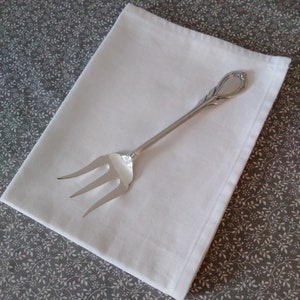 May include: A silver fork with an ornate handle, lying on a white napkin. The napkin is on a grey background with a white floral pattern.
