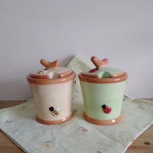 May include: Two ceramic honey pots with lids. The pot on the left is cream coloured with a bee design. The pot on the right is green with a ladybug design.