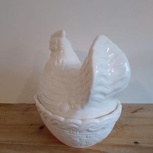 May include: A white ceramic hen-shaped container with a basket-weave base. The hen's body forms the lid, and the base is a bowl. The container is on a wooden surface.