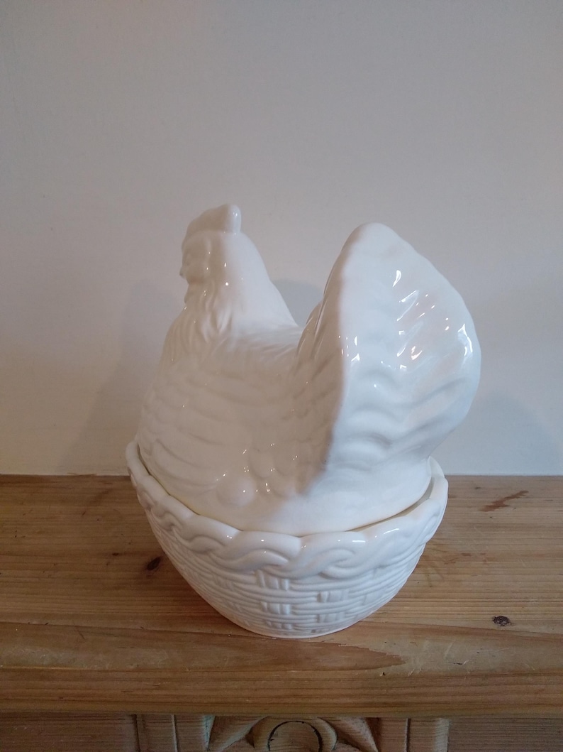 May include: A white ceramic hen-shaped container with a basket-weave base. The hen's body forms the lid, and the base is a bowl. The container is on a wooden surface.