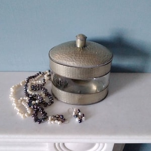 May include: A silver metal container with a hammered finish and a glass lid. The container is sitting on a white surface with a pearl necklace and earrings.