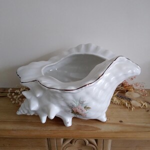 May include: White ceramic seashell planter with a gold rim and a floral design. The planter is sitting on a wooden surface.