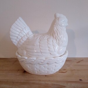 May include: A white ceramic hen-shaped container with a basket-weave base. The hen has detailed feather and facial features. The container is designed to hold eggs or other small items. The piece is sitting on a wooden surface.