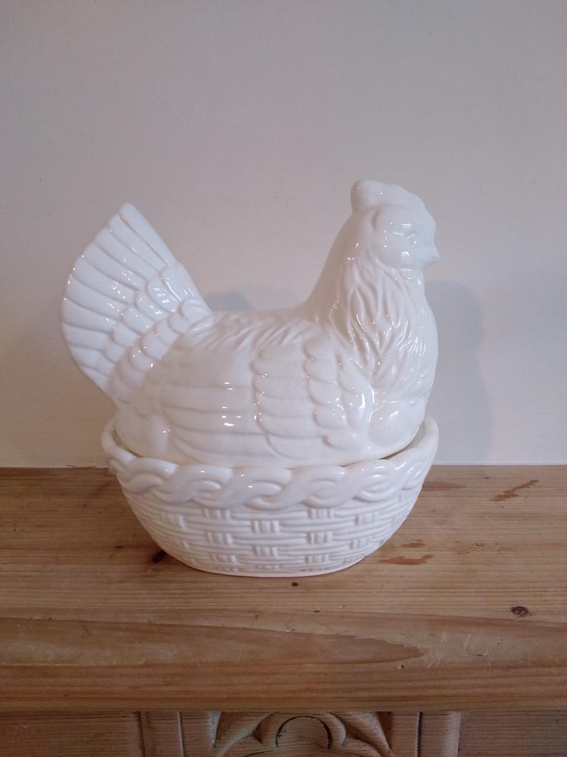 May include: A white ceramic hen-shaped container with a basket-weave base. The hen has detailed feather and facial features. The container is designed to hold eggs or other small items. The piece is sitting on a wooden surface.