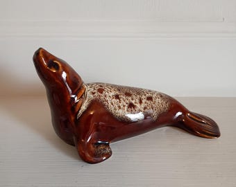 May include: A ceramic seal figurine in shades of brown and cream. The seal is posed with its head raised, showcasing a glossy, glazed finish. The body features a speckled pattern, adding texture to the decorative piece.