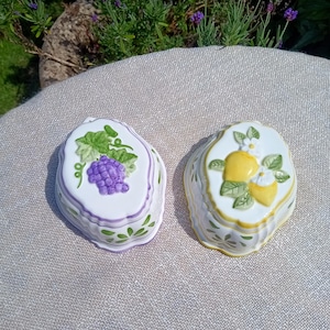 May include: Two decorative ceramic dishes. One is white with a purple grape design, green leaves, and a purple border. The other is white with a yellow lemon design, green leaves, and a yellow border. Both have scalloped edges.