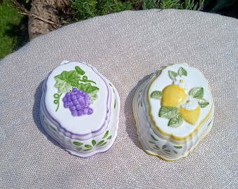 May include: Two decorative ceramic dishes. One is white with a purple grape design, green leaves, and a purple border. The other is white with a yellow lemon design, green leaves, and a yellow border. Both have scalloped edges.