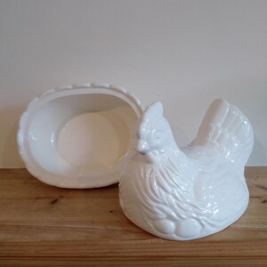 May include: A white ceramic hen-shaped dish with a matching oval-shaped lid. The hen has detailed feathering and sits on a wooden surface. The lid is open, revealing the inside of the dish. A charming kitchenware item.