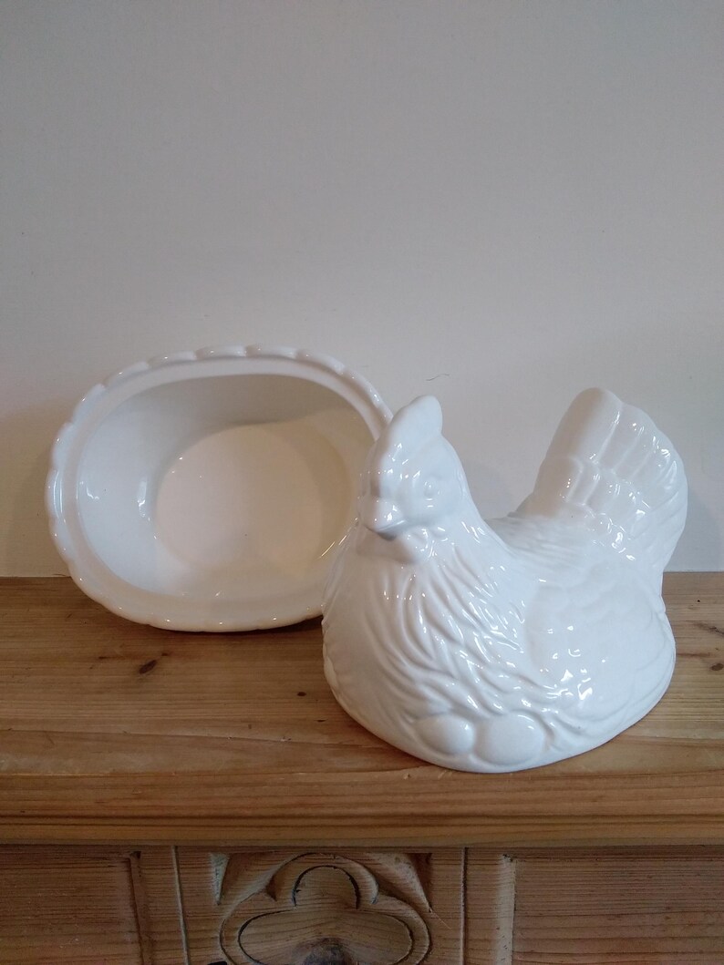 May include: A white ceramic hen-shaped dish with a matching oval-shaped lid. The hen has detailed feathering and sits on a wooden surface. The lid is open, revealing the inside of the dish. A charming kitchenware item.