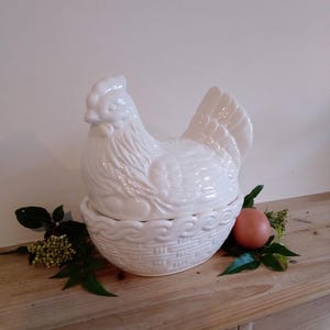 May include: A white ceramic hen-shaped container with a basket-weave base. The hen has detailed feather and facial features. An egg and greenery are next to the container on a wooden surface.