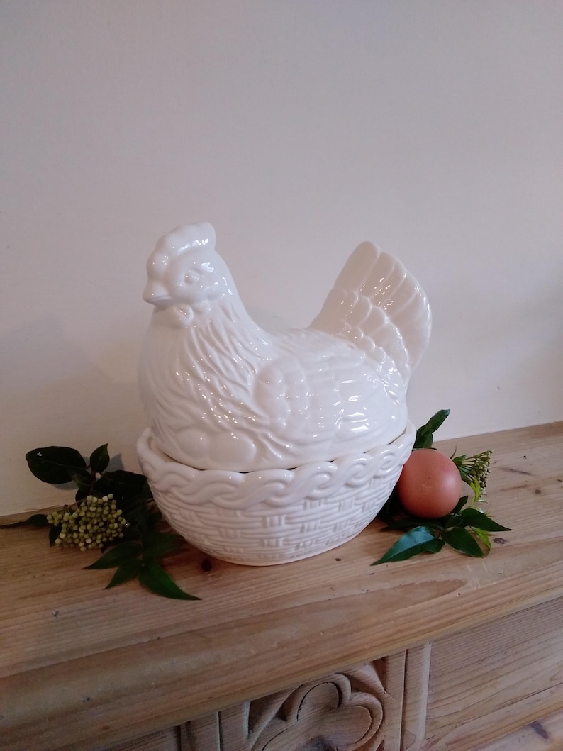 May include: A white ceramic hen-shaped container with a basket-weave base. The hen has detailed feather and facial features. An egg and greenery are next to the container on a wooden surface.