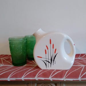 May include: A white ceramic jug with a black and red cattail design, beside two green patterned drinking glasses. The jug is on a red and white patterned tablecloth, with a wooden surface beneath.