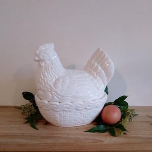 May include: A white ceramic hen-shaped egg holder with a basket-weave base. The hen is on a bed of greenery, with a single brown egg. The piece is displayed on a wooden surface. A decorative kitchen item.